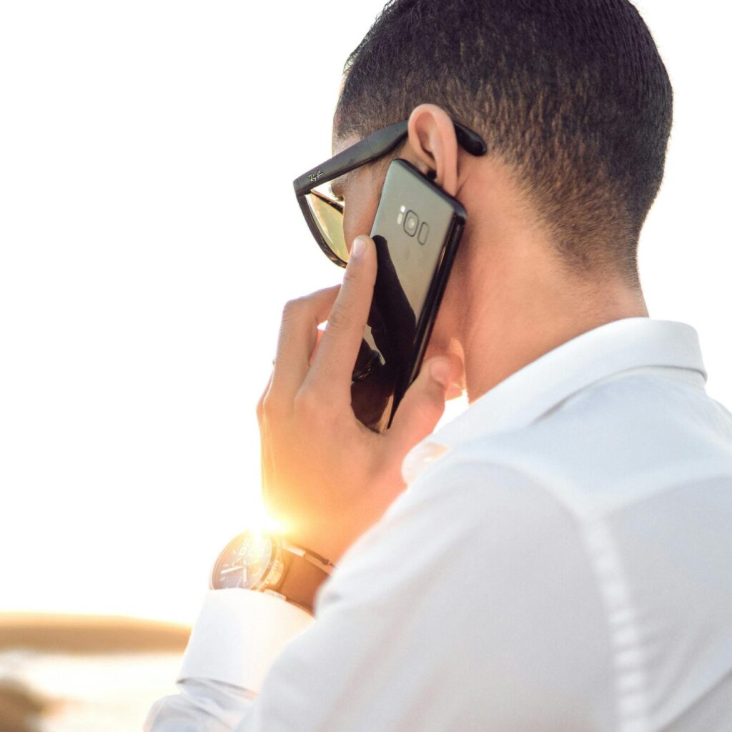 Man outdoors in Morocco talking on smartphone, wearing sunglasses and white shirt, backlit by sunset.